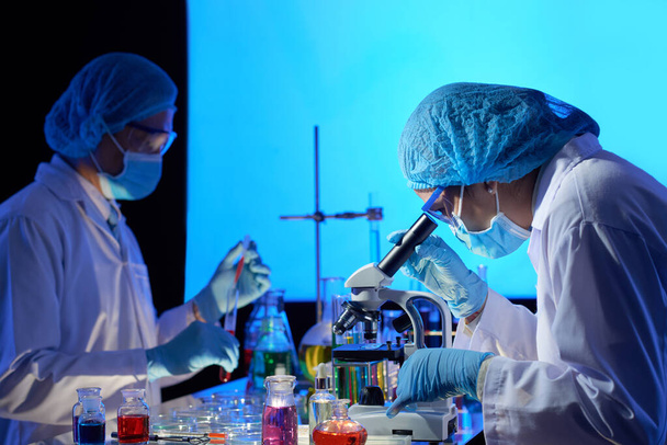 Equipe de cientistas trabalhando na vacina covid-19 em laboratório escuro, misturando líquidos e olhando através do microscópio
 - Foto, Imagem
