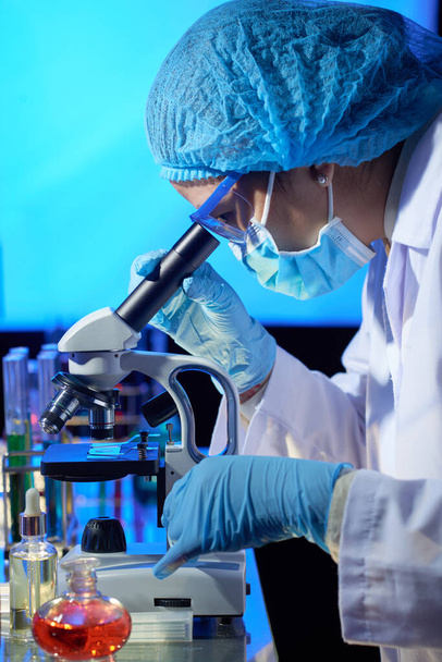 Laboratorion työntekijä lääketieteellisessä hatussa, naamiossa ja käsineissä mikroskoopilla - Valokuva, kuva