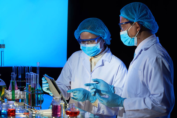 Techniciens de laboratoire en masques médicaux discutant des détails de recherche sur tablette avant de mélanger des réactifs - Photo, image