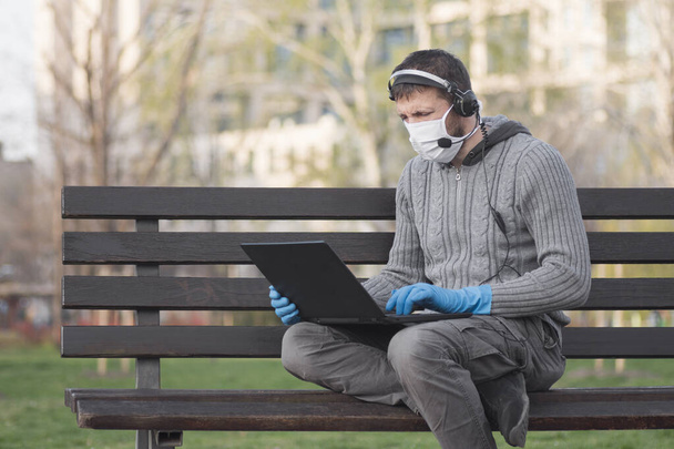 Mies suojaava naamio ja käsineet kuulokkeella käyttää kannettavaa ulkona, istuu penkillä puistossa. - Valokuva, kuva