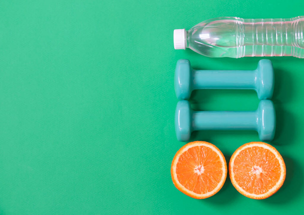 Mancuernas azules, botella de agua y naranjas como borde sobre fondo verde. Concepto de estilo de vida saludable. Vista superior, espacio de copia
. - Foto, imagen