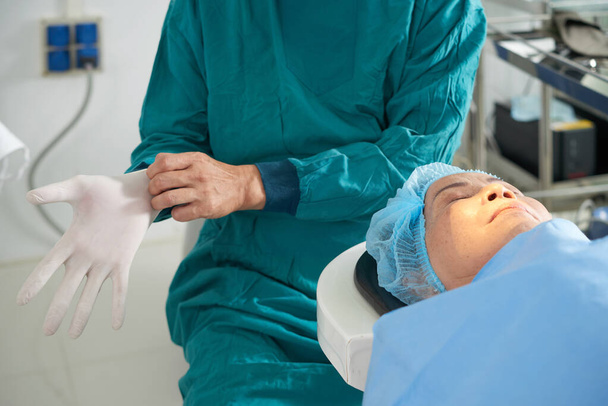 Старшая женщина лежит на операционном столе в офтальмологической клинике с закрытыми глазами, хирург надевает резиновые перчатки - Фото, изображение