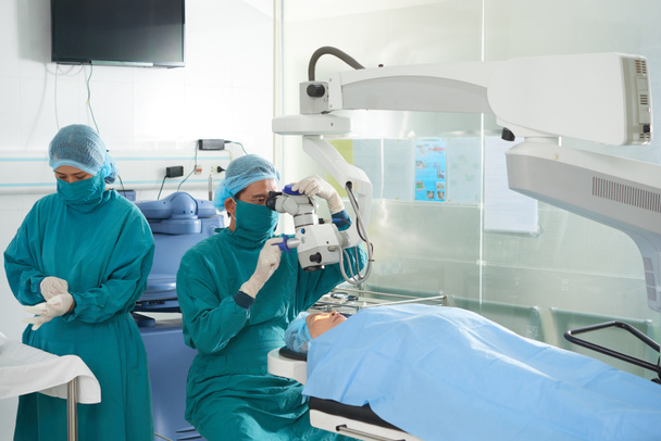 Cirujano que ajusta el láser oftálmico sobre la cabeza del paciente cuando el asistente se pone guantes de goma - Foto, Imagen
