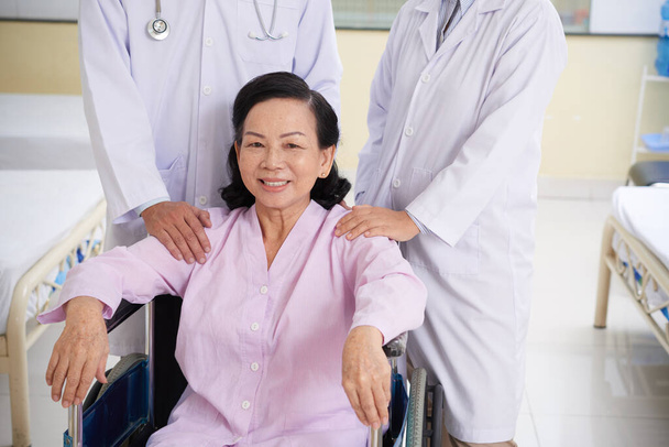 Счастливая азиатская старшая женщина, сидящая в инвалидной коляске после лазерной хирургии глаза с врачами, стоящими рядом - Фото, изображение