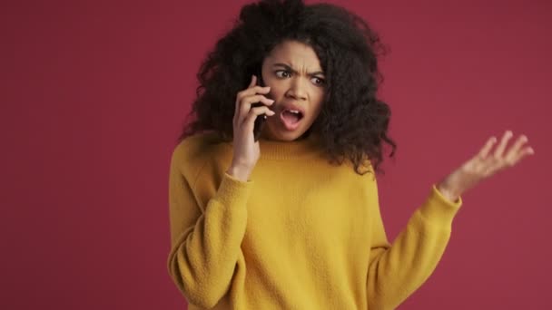 Young african confused displeased woman isolated over dark red burgundy background talking by mobile phone - Кадры, видео
