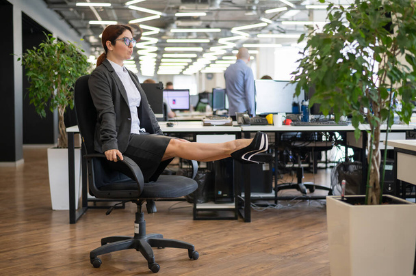 Happy business woman doing abdominal muscle exercises in an open space office. A red-haired smiling female employee in a skirt and high heels is dying off a chair in the workplace. Fitness at work. - Photo, image