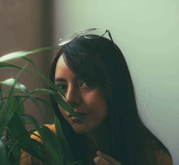 Hispanic girl looking towards the camera, covered by the leaves of a bamboo plant, old photo, the girl wears a yellow blouse and wears magnifying glasses on her head, lifestyle photography - Foto, Imagem