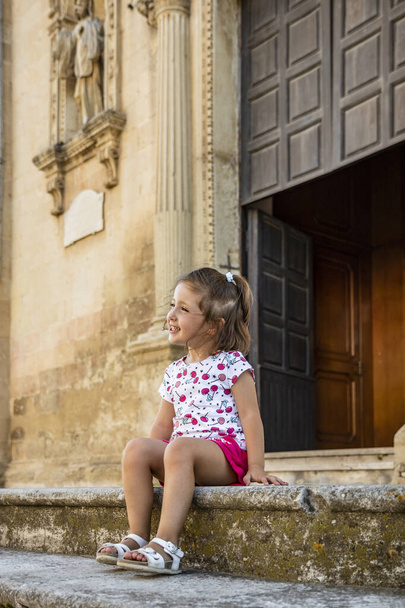 Kaunis ja hymyilevä pikkutyttö, joka istuu Cavallinon, Leccen, Puglian, Salenton, Italian barokkikirkon portailla. Söpö lapsi hymyilee poseeratessaan valokuvaa varten. Puinen portaali taustalla. - Valokuva, kuva