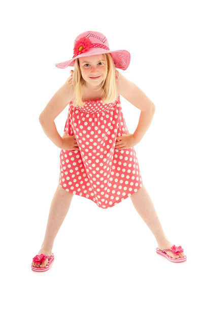 ピンクのフロッピー帽子、ポルカドットドレス、フリップフロップを身に着けている若いブロンドの女の子の完全な長さのショット。白いスタジオの背景に隔離された - 写真・画像