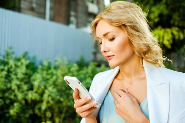 Nahaufnahme Außenporträt einer hübschen Geschäftsfrau mit Smartphone. Model hat blonde Haare und trägt schönen weißen Anzug. - Foto, Bild