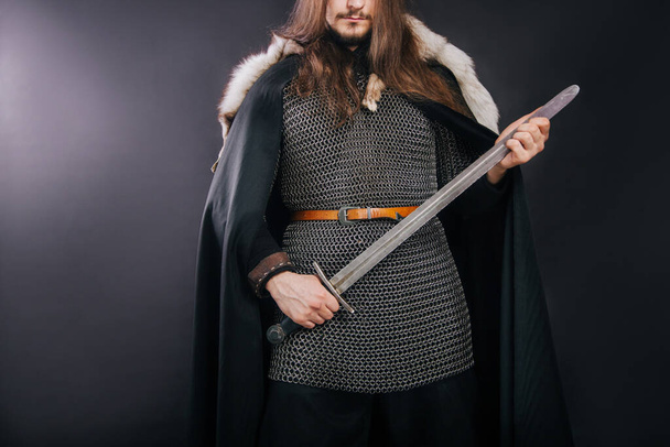 Knight in chain mail and with a fur collar on a black cloak on a gray background. Portrait of a Viking man with long hair and a beard in armor holds a sword. - Foto, Imagem
