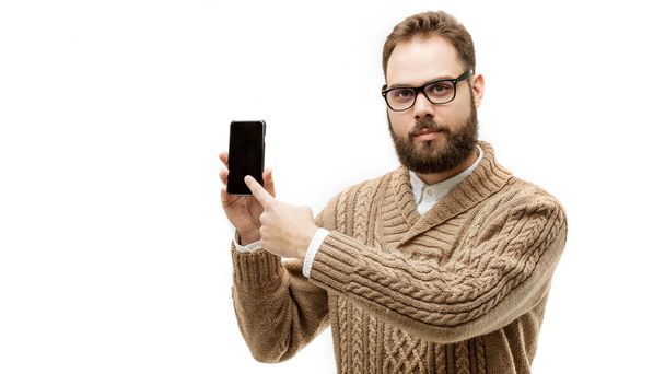 closeup portrait of handsome man with beautiful beard wearing eyeglasses and warm brown sweater. Model is pointing index finger on touch screen of his smartphone isolated on white background - Photo, Image