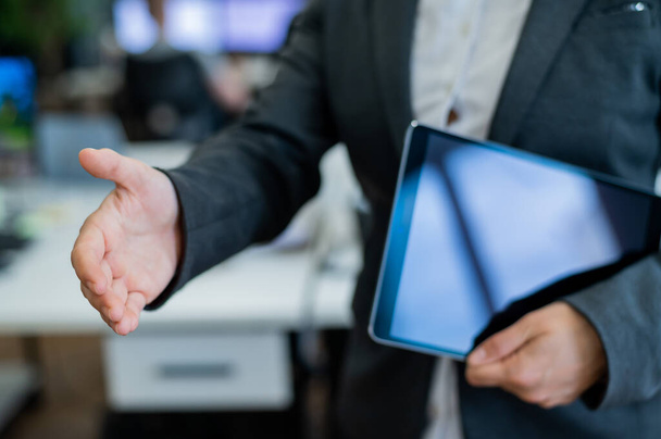 Business woman in a jacket and white shirt holding out a hand for a greeting. A faceless business woman holds a digital tablet and shakes her hand as an agreement on a successful transaction. - Фото, изображение