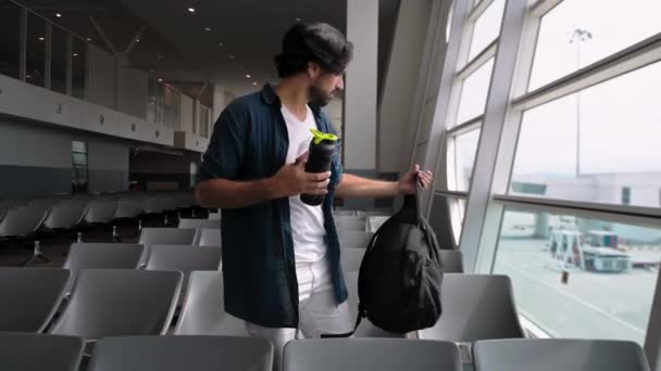 De jonge, knappe man zit alleen op het vliegveld in een wachtkamer. Een knappe vent kijkt naar zijn paspoort en ticket. Positieve toerist op de luchthaven voor een vlucht. - Video