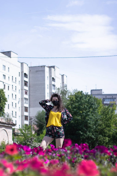 Attractive young brunette woman in yellow t-shirt and flowery shorts costume. In medical black face mask. modern reality. near bushes with flowers. covid-19 concept. copy space. - Фото, зображення