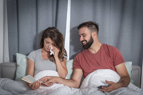Young man making fun and laughing because his girlfriend crying while she reading a book in the bed and cry - Фото, изображение