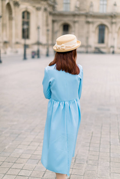 Back view of beautiful elegant young lady in hat and blue dress, walking near the vintage building in European city - Fotografie, Obrázek