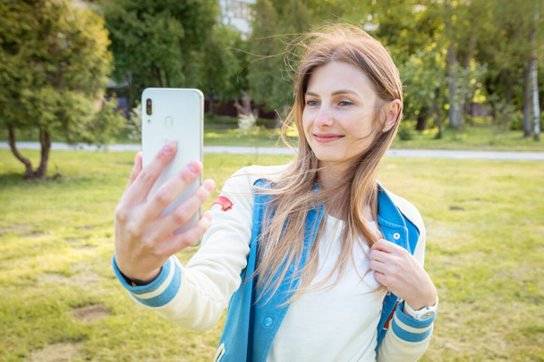 Joven hermosa chica toma fotos de sí misma con un teléfono inteligente. Buen tiempo de verano. La mujer está en el parque, la calle es soleada y cálida
.  - Foto, imagen