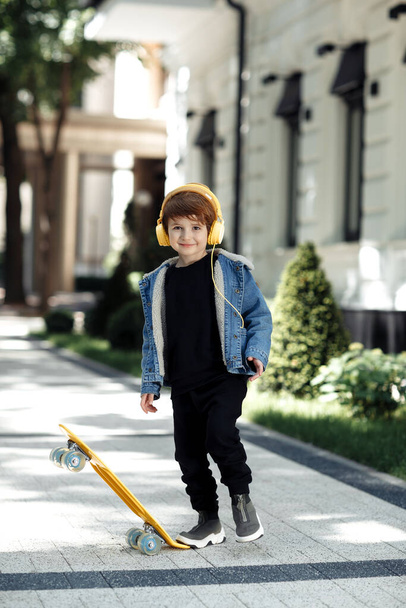 Photo of joyful little boy posing with headphones and skateboard in street. - Zdjęcie, obraz