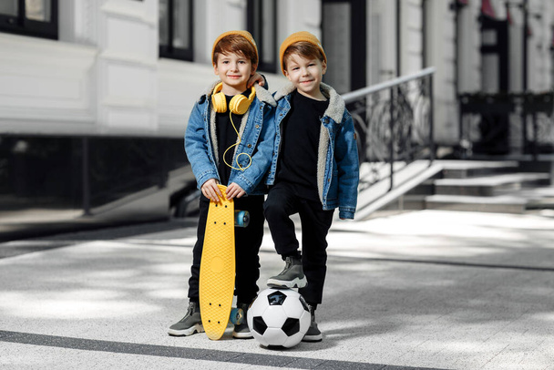 Two funny twin boys posing with happy faces in the street. Skateboard or pennyboard and soccer ball. - Photo, image