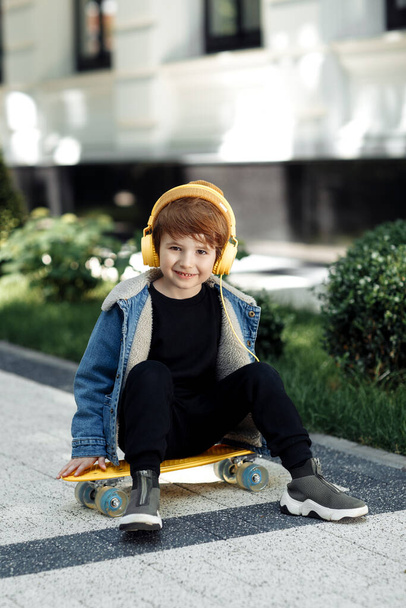 Photo of cute little boy sitting on the skateboard or pennyboard listening music in headphones in street. - Photo, image