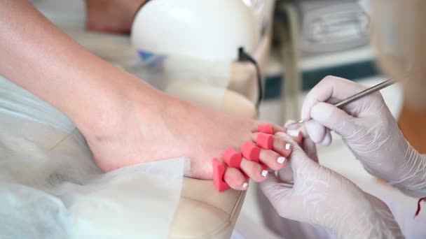 シェラックランプとつま先の指のセパレーターを使用して、クライアントの足に白いネイルポリッシュを行うペディキュリスト。専門的な医療画像処理手順.SPAサロンでの足の治療.内科診療所。手袋をして手. - 映像、動画