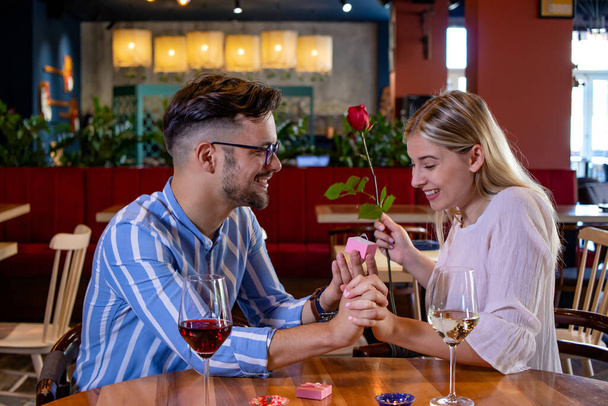 Jong romantisch stel in een mooi chique restaurant. Vriendje verrassen zijn vriendin met een rode roos en haar ten huwelijk vragen met een verlovingsring. - Foto, afbeelding