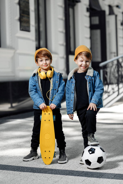 Two cheerful twin boys posing with happy faces in the street. Skateboard or pennyboard and soccer ball. - 写真・画像