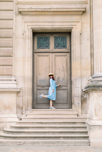 Charming elegant young woman wearing blue dress, posing in front of ancient wooden door of old vintage building with one leg up - Foto, Bild