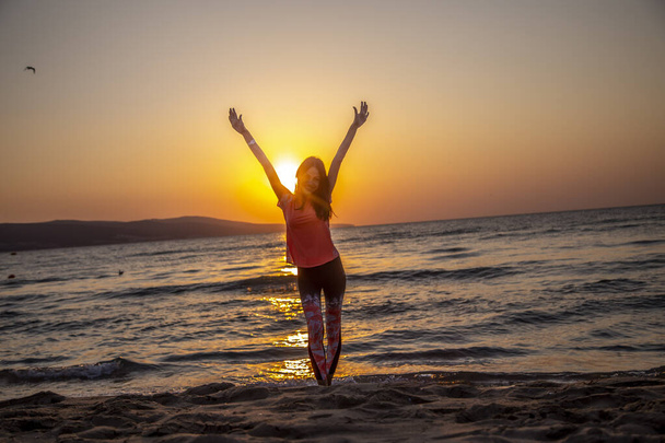 Mulher feliz em pé na praia de areia com as mãos para cima, braços abertos no belo nascer do sol à beira-mar
 - Foto, Imagem