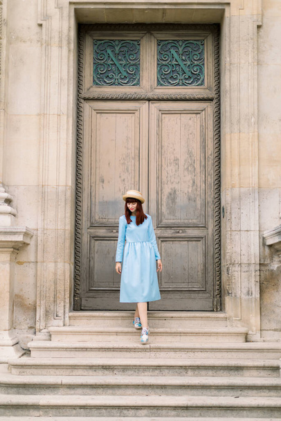 Photo of a beautiful young woman in blue dress descends the vintage stairs on the background of old building with doors - Foto, Imagem