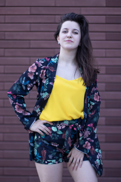 Mulher morena jovem atraente em camiseta amarela e traje de shorts floridos. fundo da parede marrom. espaço de cópia
. - Foto, Imagem