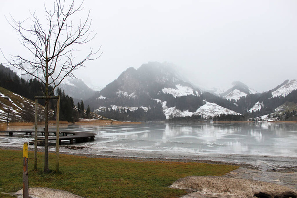 Schwarzsee of Lac Noir is een meer in het kanton Friborg in Zwitserland. Houten steiger op het bevroren meer, ondergedompeld in de mist, tussen de besneeuwde bergen - Foto, afbeelding