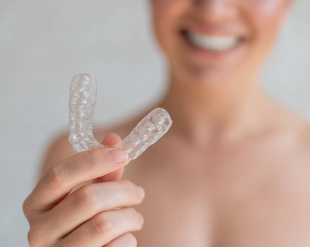 Close-up of orthodontic silicone transparent teeth aligner in female hands. Blurred unrecognizable woman holds a removable night retainer. Bracket for teeth whitening for a perfect smile. - Фото, изображение