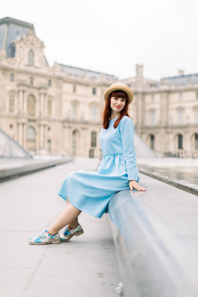 PARIS, FRANCE - September 17, 2019: Young happy woman in blue dress, sitting near the fountain and posing for a photo with the glass pyramid background at Louvre museum - Zdjęcie, obraz