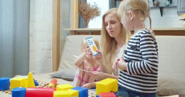 Young mother showing two daughters cards with letters and pictures studying the English alphabet while sitting on the sofa at home. Preschool education. - Imágenes, Vídeo