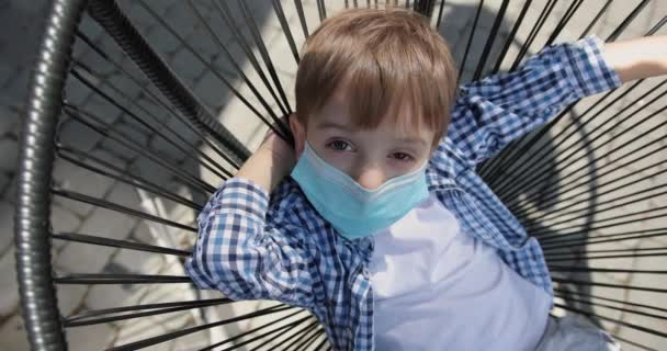 Kleine jongen met een medisch masker met pijnlijke ogen op een stoel. Coronavirusepidemie. - Video