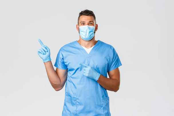Healthcare workers, covid-19, coronavirus and preventing virus concept. Happy professional male nurse, doctor in scrubs and medical mask, gloves pointing finger left, showing banner - Photo, Image