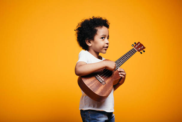 Black-skinned curly boy in a white T-shirt plays ukulele guitar and looks at the camera on an orange background in the studio - Photo, Image