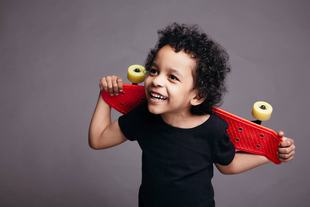 Close-up of a curly dark skinned boy in a black T-shirt holding red skateboard behind his back and smiling on gray background - Foto, afbeelding