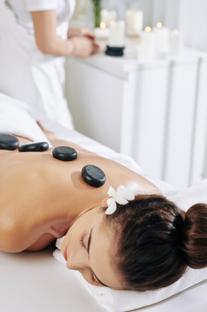 Junge Frau mit Blume im Haar bekommt Hot Stone Massage im Wellness-Salon - Foto, Bild