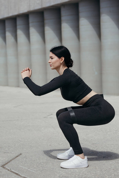 Close up of a fitness trainer in a black sports uniform doing squats on the street against the background of concrete cylinders. - 写真・画像