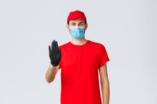 Covid-19, self-quarantine, online shopping concept. Confident delivery man in red uniform, gloves and face mask, raise hand in stop sign, prevent client step to storage without protective equipment - Photo, Image