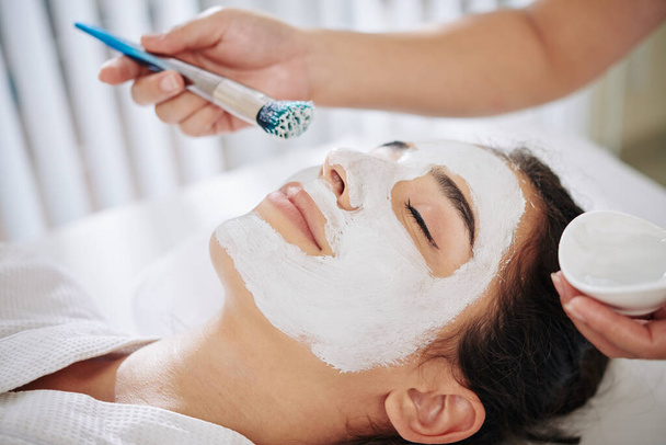 Cosmetólogo usando cepillo al aplicar máscara de arcilla blanca en la cara del cliente
 - Foto, Imagen