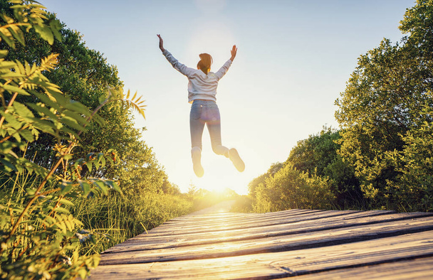 Mutlu kadın kaldırılmış eller ve bacaklarla atlıyor ve günbatımında tahta bir yolda hayatın tadını çıkarıyor. - Fotoğraf, Görsel