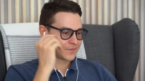 Young handsome brunette man sits in gray chair, listens to music on smartphone, keep calm and relax. Caucasian happy guy wears black glasses, white headphones, blue polo shirt, close up. Copy space. - Footage, Video