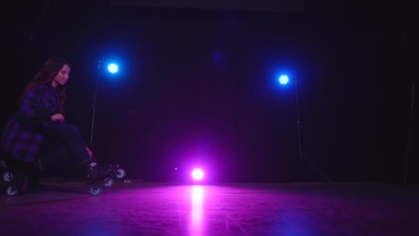 Avvicinamento vista di luce rosa di riflettori tra lampade blu e una ragazza pattinaggio a rotelle seduto su ruote anteriori e posteriori sul palco buio. Concetto di sport e stile di vita sano. Casting in studio. - Filmati, video