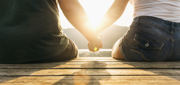 Joyeux moments ensemble. Heureux jeune couple tenant la main assis sur la jetée au bord d'un lac au coucher du soleil
 - Photo, image