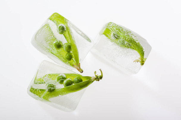 Draufsicht auf frische grüne Erbsenschoten eingefroren in Eiswürfeln auf weißem Hintergrund. Konzept des Einfrierens von Früchten - Foto, Bild