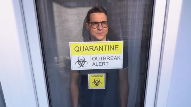 Worried Isolated infected sick young man wearing black glasses, T-shirt leans glass, looks out from hospital room close up. Yellow white sign inscription on window Quarantine Outbreak Alert. Lock down - Footage, Video